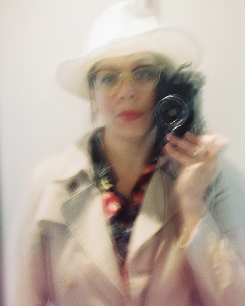 Auto portrait de Oleñka Carrasco. Elle tient l'appareil photo contre sa joue gauche. Elle est habillée avec un trench beige, dont on voit dépasser un chemisier à fleur. Elle porte un large chapeau blanc, et de grandes lunettes.