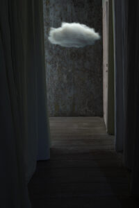 Cadrage vertical. Intérieur nuit. Lumière et couleur bleutées de pleine lune. Au bout de la perspective d’un court et obscur couloir bordé de part et d’autre de rideaux, un petit nuage clair se détache à un mètre cinquante (tiers supérieur du cadre) du sol en parquet sur le fond d’un mur sombre à la texture marbré. Dans le prolongement du mur droit du couloir, une porte fermée de bois peint en blanc cassé retient le petit nuage de s’échapper.