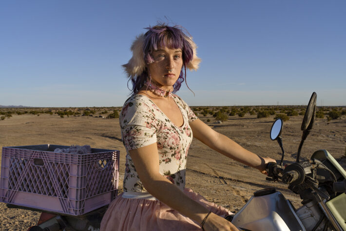Une femme à cheveux violets et blancs sur une moto dans le désert.