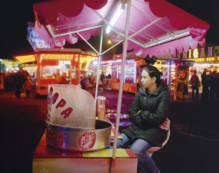 Une femme assise sous un parasol rose à un stand de barbe à papa dans une fête foraine illuminée la nuit.