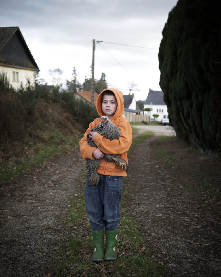 Un enfant en orange tenant un poulet dans un chemin rural.