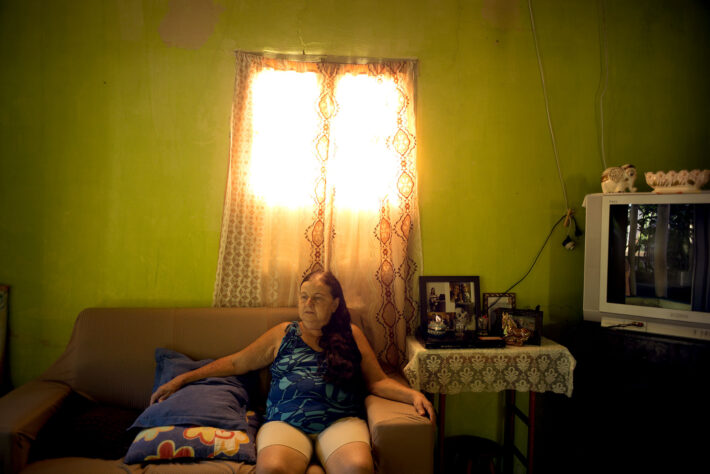 Femme assise sur un canapé dans un salon à la lumière dorée, avec des photos de famille et une télévision ancienne à côté d’elle.