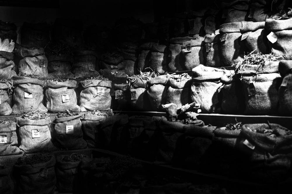 La photographie, en noir et blanc, montre la réserve de plantes d’une herboristerie familiale au Vietnam. Le premier plan et l’arrière-plan de la photographie sont sombres, ce qui fait ressortir, par contraste, les sacs de toile contenant les herbes médicinales, rangés sur des étagères et étiquetés. Les sacs sont tous ouverts laissant apparaître les plantes qu’ils contiennent.